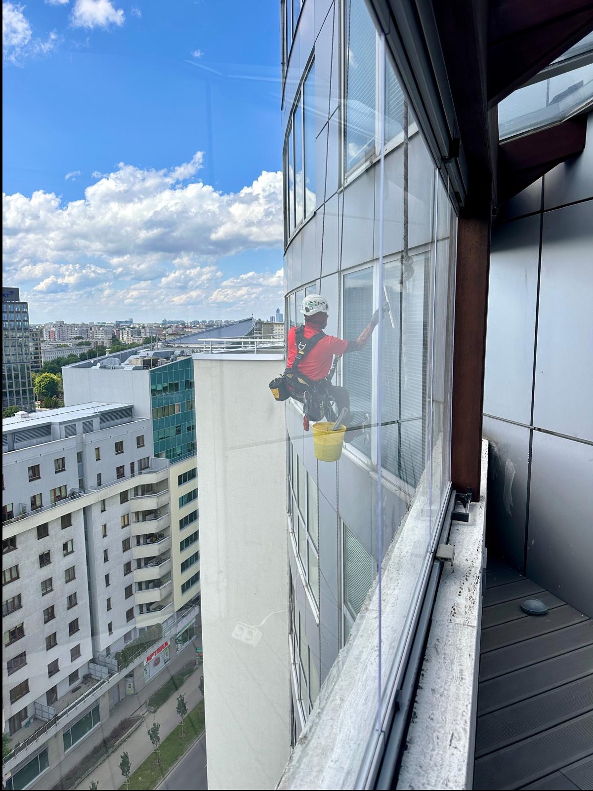 Pracownik w kasku i uprzęży, zawieszony na linie, czyści okna wieżowca za pomocą ściągaczki, widok na miasto w tle.