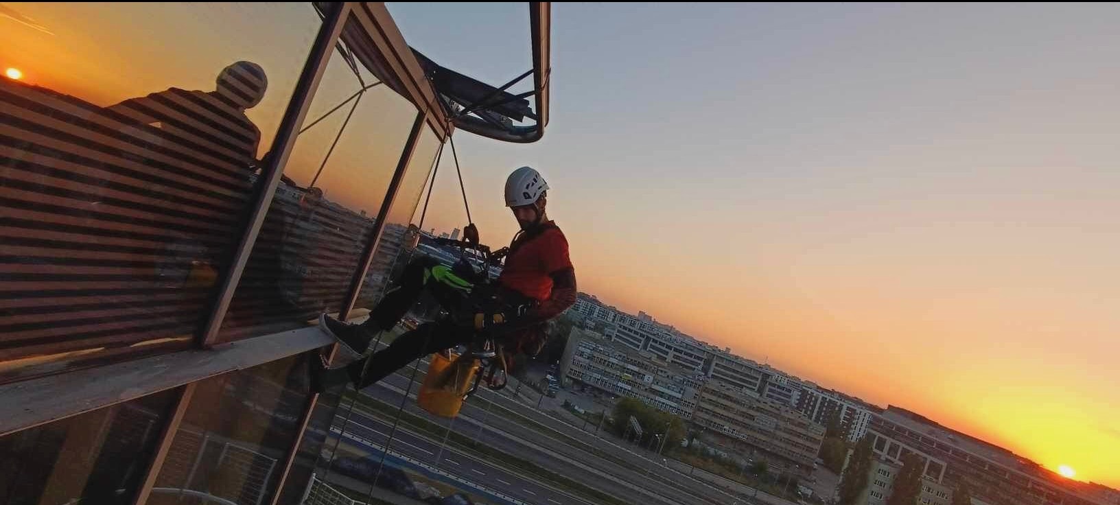 Pracownik w kasku i uprzęży, zawieszony na linach, czyści szklaną fasadę budynku na wysokości, z widokiem na zachodzące słońce i panoramę miasta.