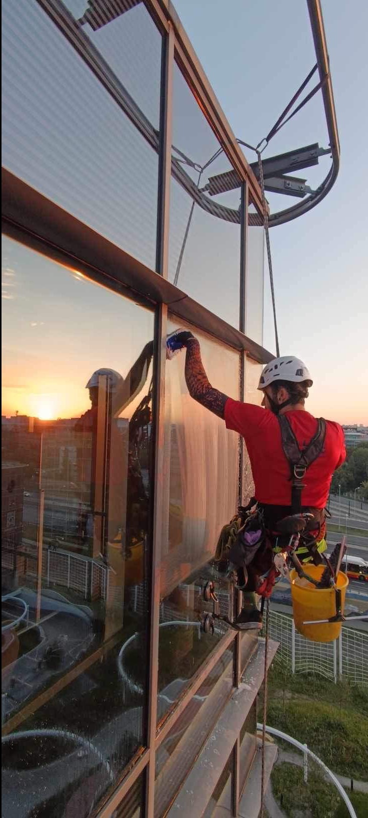 Pracownik w uprzęży, z tatuażem na ramieniu, czyści okna na wysokości w budynku biurowym, w tle zachód słońca i ruchliwa ulica.