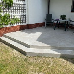 ART BUD ARTUR KAVALEVICH - Wyłożony płytkami taras z dwoma stopniami, z widoczną kratką na pnącza na ścianie budynku, stolik z rośliną doniczkową i krzesła w tle.