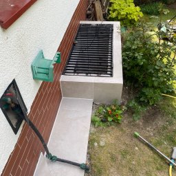 ART BUD ARTUR KAVALEVICH - Murowany grill przy ścianie domu, wykończony jasnymi płytkami, z widocznymi elementami instalacji wodnej i zielenią w tle.