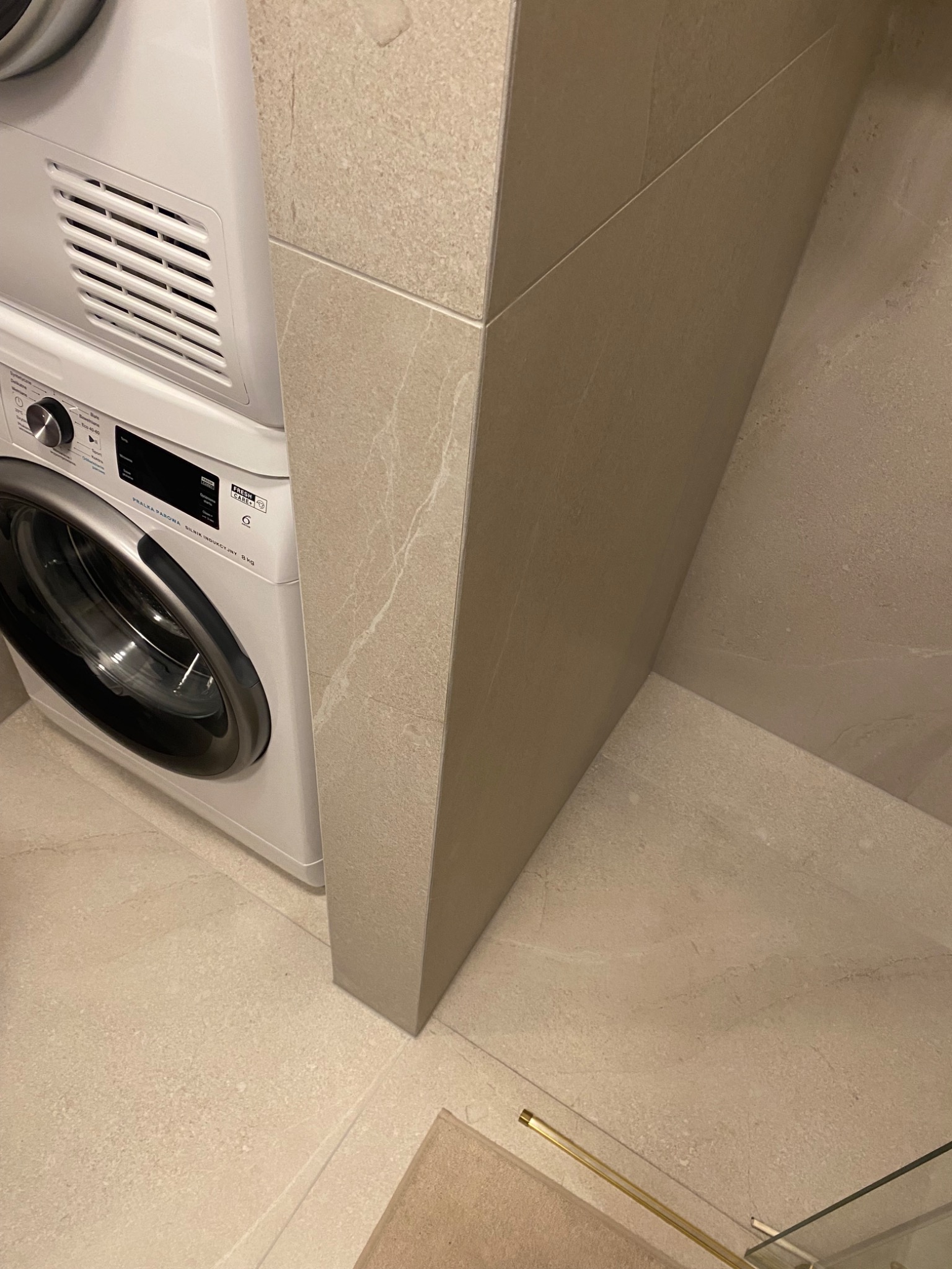Fragment łazienki z pralko-suszarką w zabudowie, wykończonej płytkami w odcieniach beżu z delikatnym wzorem marmuru.