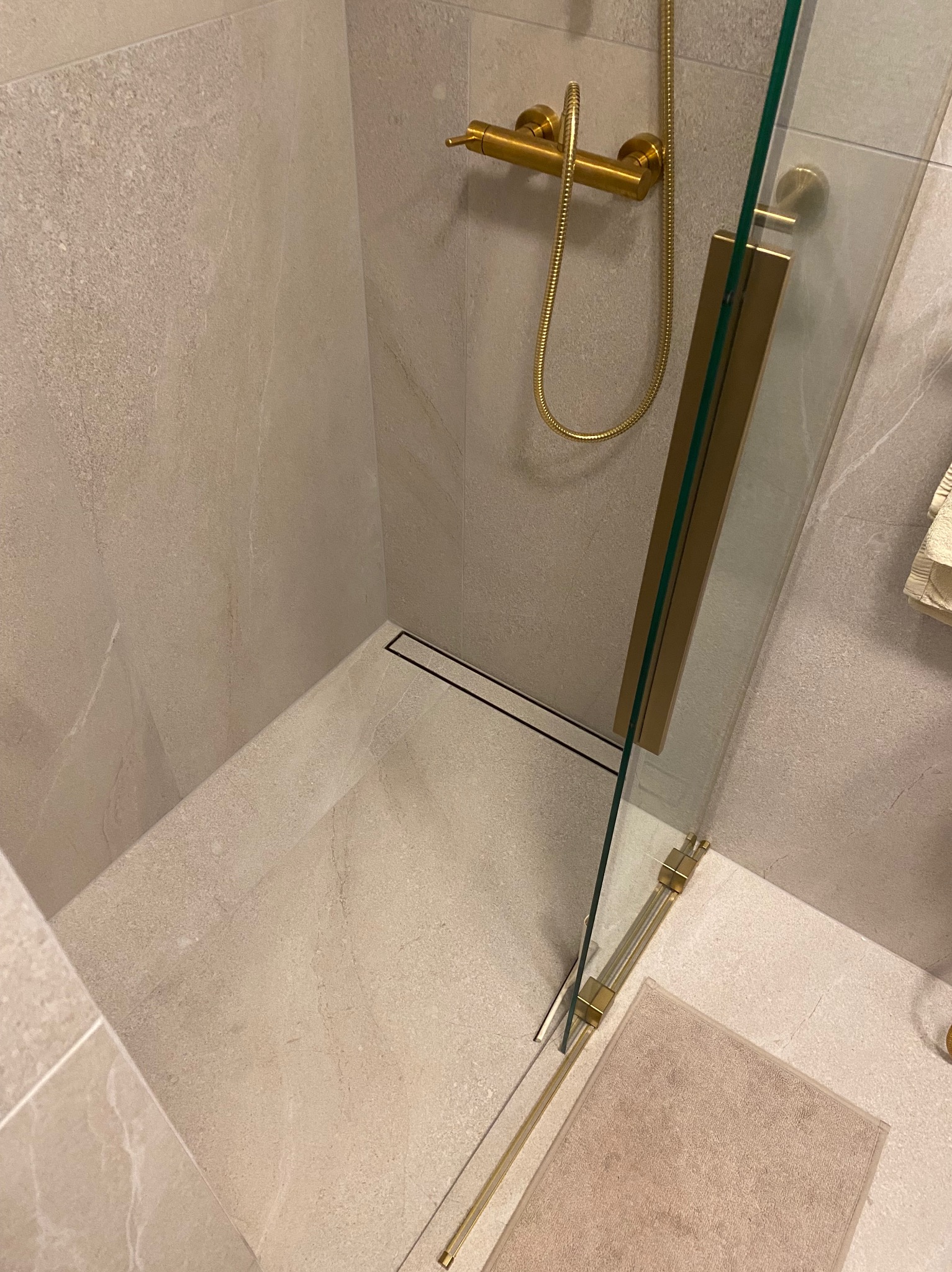 Beżowa kabina prysznicowa z odpływem liniowym, złotą armaturą i szklanymi drzwiami, wyłożona płytkami imitującymi kamień.