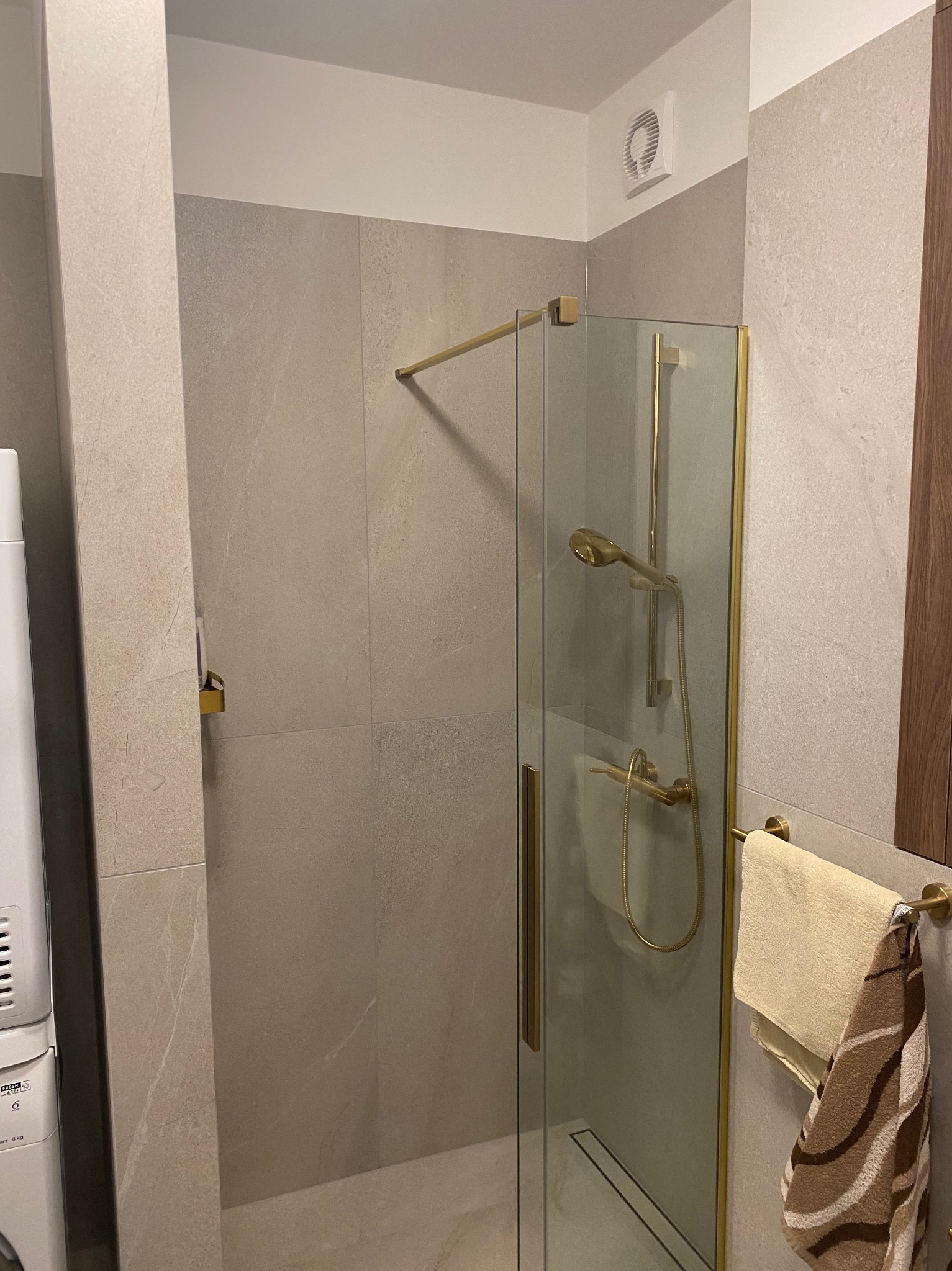 Wykończona łazienka z kabiną prysznicową w złotej ramie, beżowe płytki imitujące kamień, widoczny fragment pralki i ręczniki na złotym wieszaku.