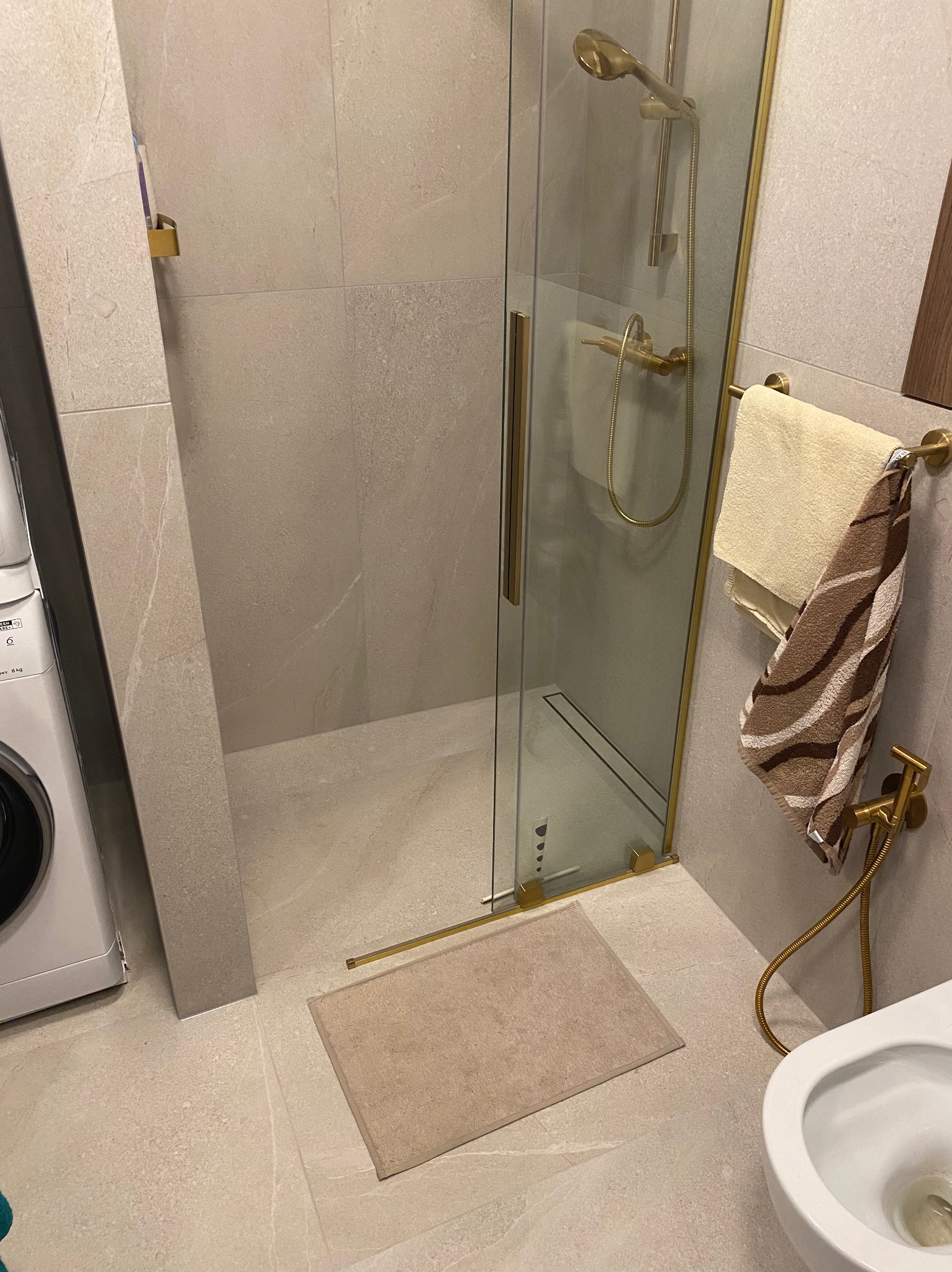 Nowoczesna łazienka z kabiną prysznicową w odcieniach beżu i złota, widoczna pralka i dywanik pod prysznicem.