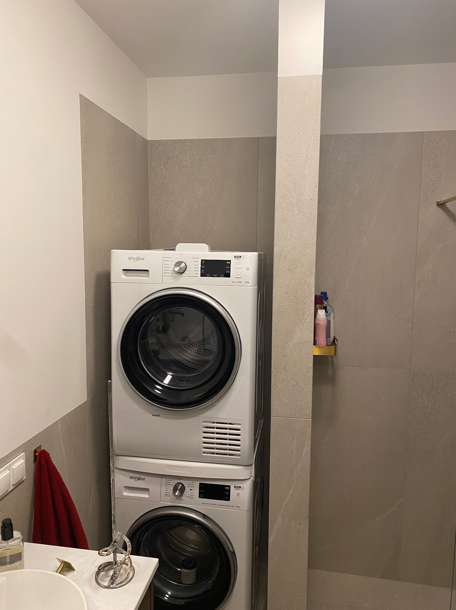 Ustawiona w pionie pralka i suszarka Whirlpool w łazience wyłożonej szarymi płytkami, obok umywalka z akcesoriami i czerwony ręcznik.