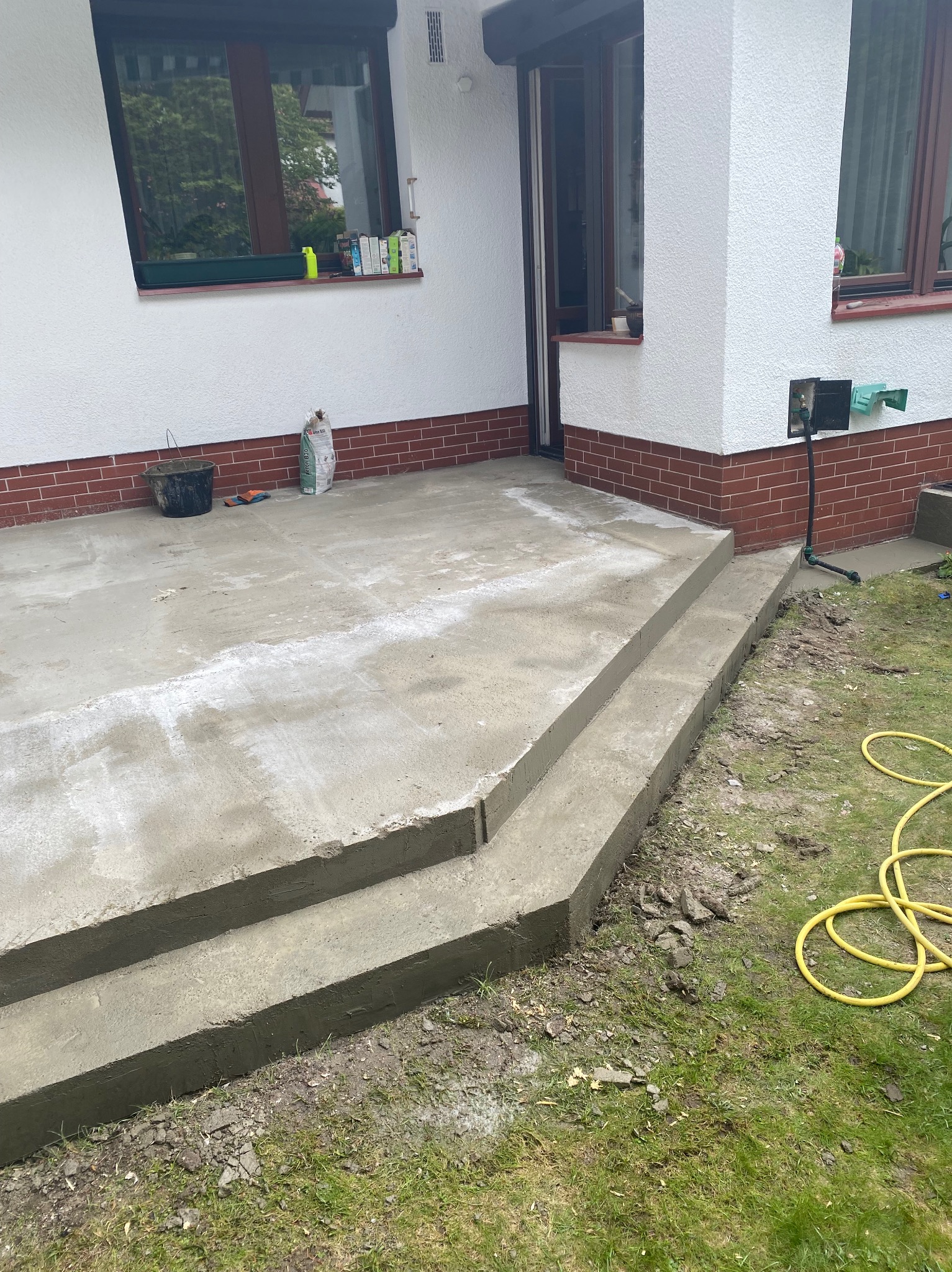 Świeżo wylany betonowy taras z dwustopniowymi schodami przy budynku z ceglaną podmurówką, widoczne narzędzia i worek z zaprawą.