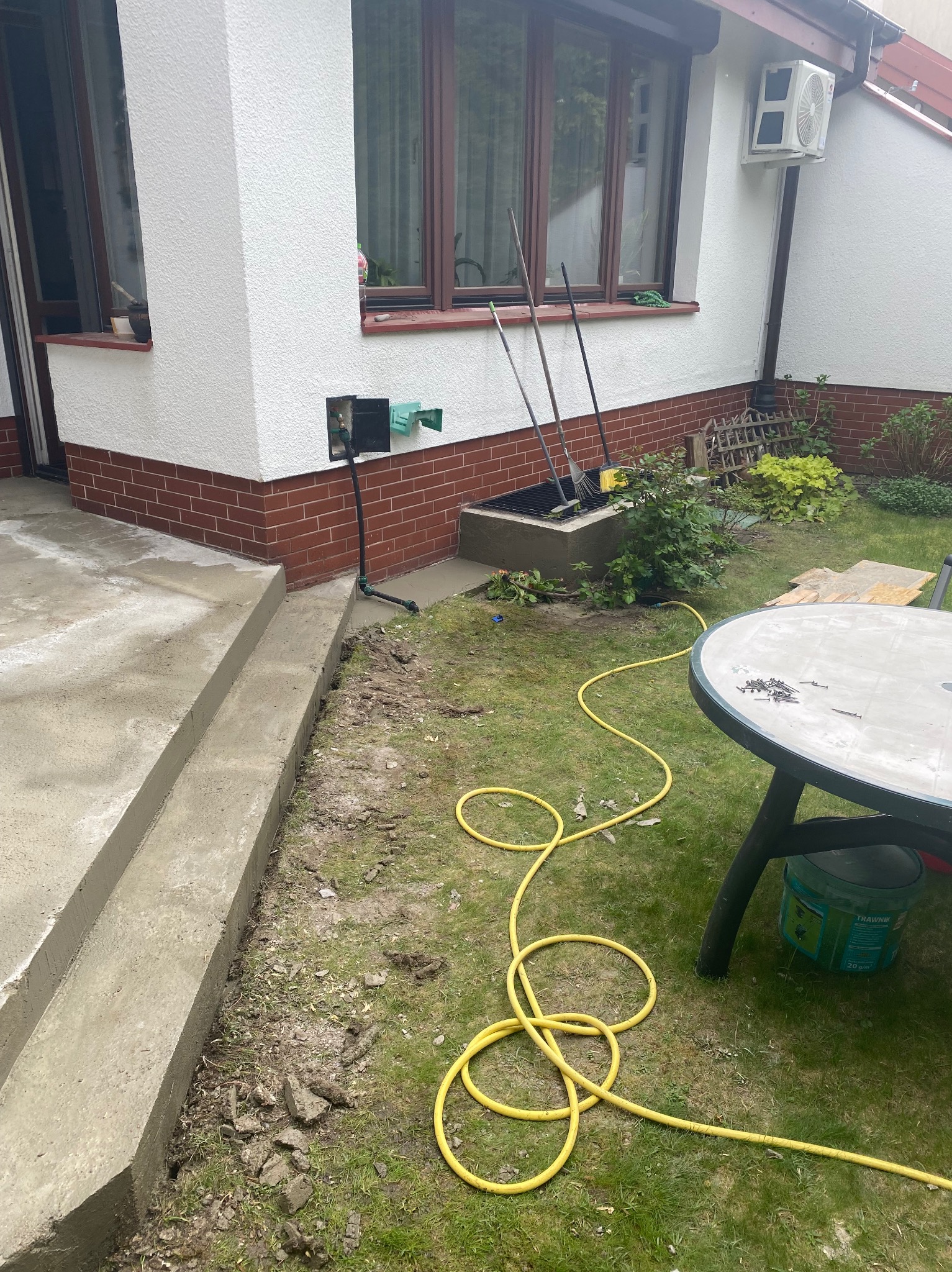 Świeżo wylane betonowe schody zewnętrzne przy domu z ceglaną podmurówką, żółty wąż ogrodowy na trawie, widoczne narzędzia i stół ogrodowy z gwoździami.