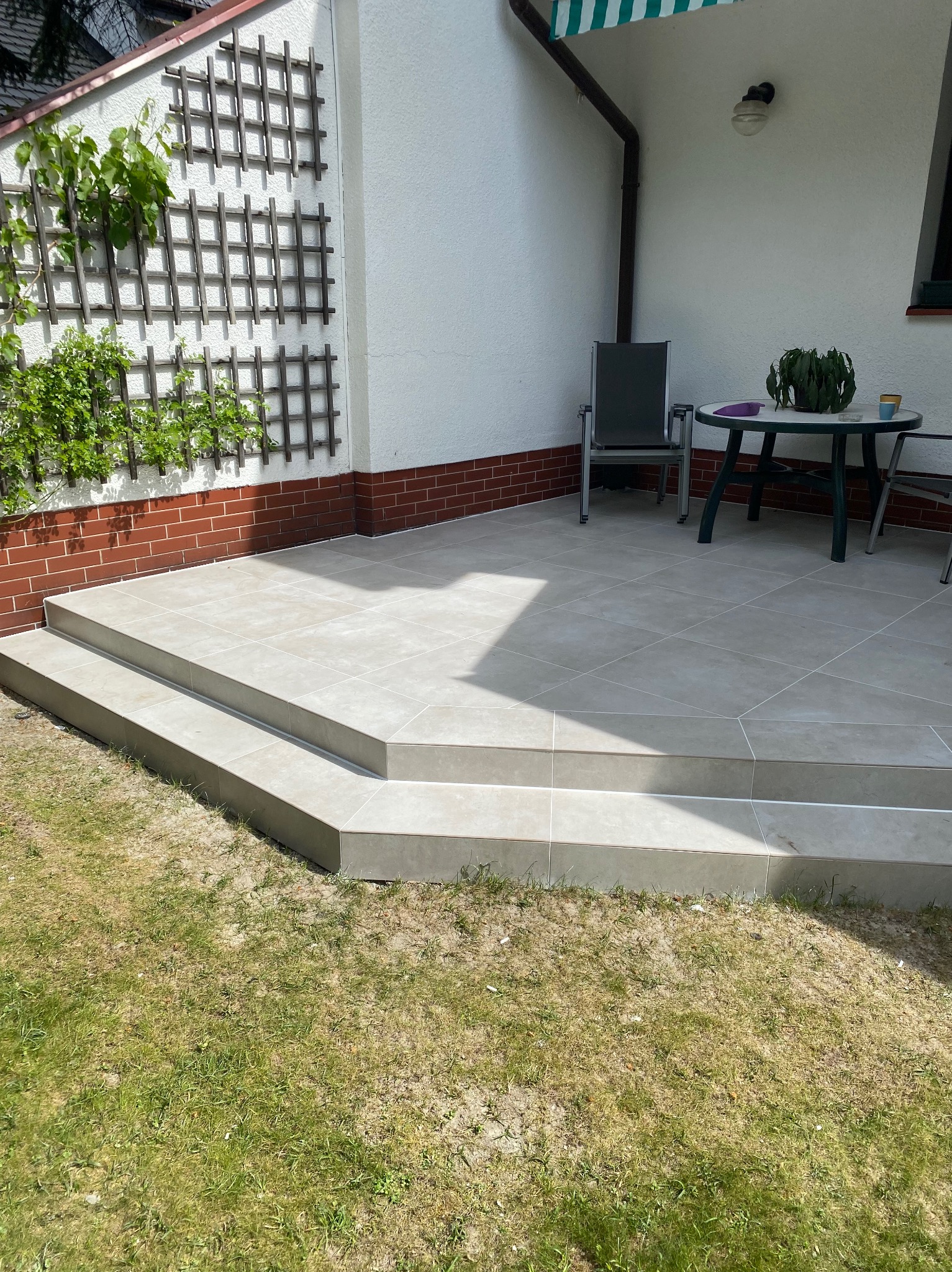 Wyłożony płytkami taras z dwoma stopniami, z widoczną kratką na pnącza na ścianie budynku, stolik z rośliną doniczkową i krzesła w tle.