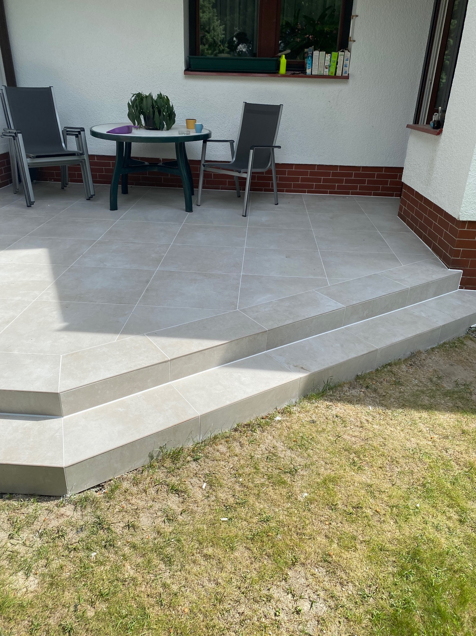 Wyłożony jasnymi płytkami taras z dwoma stopniami, widoczne krzesła i stolik z rośliną doniczkową