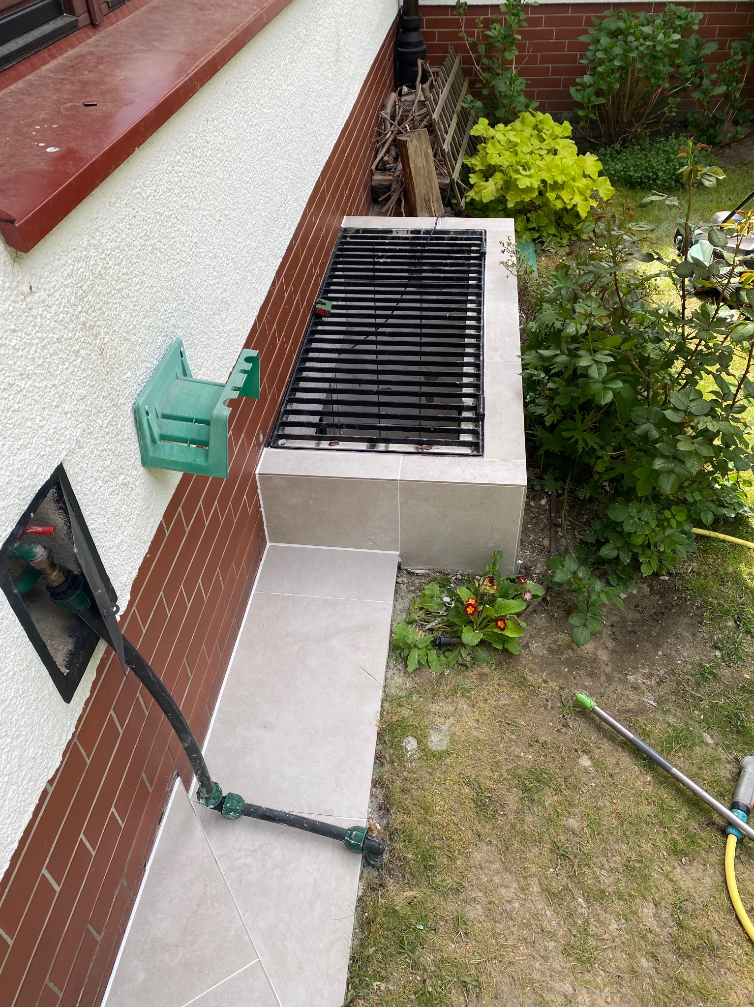 Murowany grill przy ścianie domu, wykończony jasnymi płytkami, z widocznymi elementami instalacji wodnej i zielenią w tle.