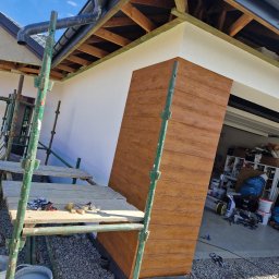 Tom-bud - Montaż paneli elewacyjnych imitujących drewno, widoczne rusztowanie, fragment elewacji w kolorze białym i otwarte drzwi garażowe z widocznym bałaganem w środku.