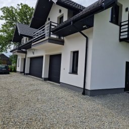 Tom-bud - Nowoczesny, biały budynek z trzema garażami i balkonami z czarnymi elementami wykończeniowymi, otoczony żwirowym podjazdem i zielenią.