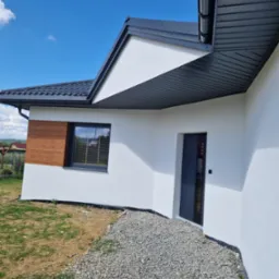 Nowoczesny dom z białą elewacją, drewnianym akcentem obok okna i szarą stolarką okienną i drzwiową, otoczony kamiennym podłożem i trawnikiem w słoneczny dzień.