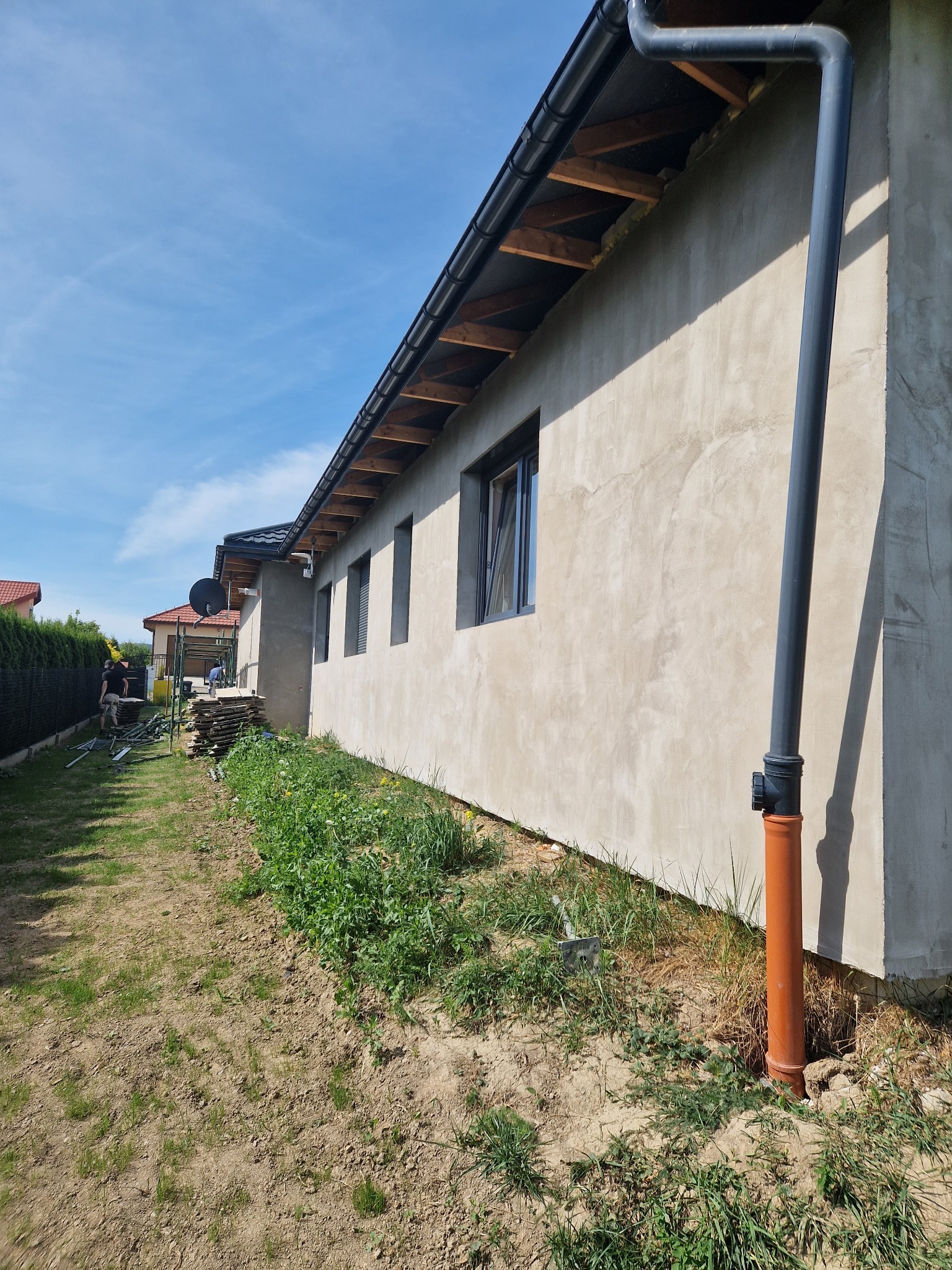 Surowa elewacja domu jednorodzinnego z widocznymi oknami i rurą spustową, częściowo wykończona, z elementami rusztowania i składowanym materiałem budowlanym w tle, w słoneczny dzień.