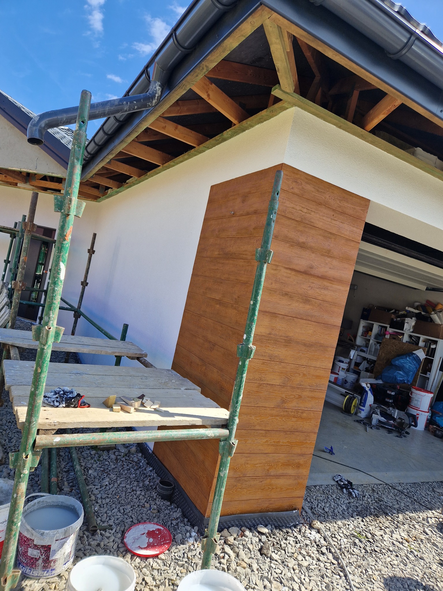 Montaż paneli elewacyjnych imitujących drewno, widoczne rusztowanie, fragment elewacji w kolorze białym i otwarte drzwi garażowe z widocznym bałaganem w środku.