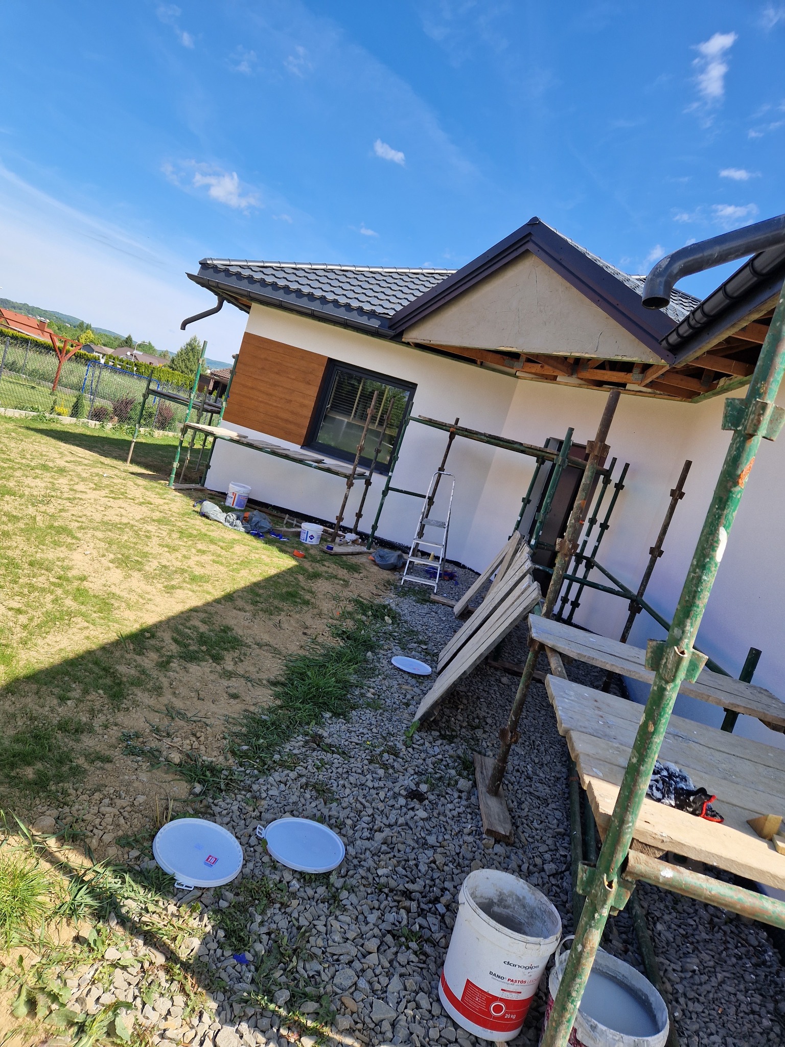 Dom jednorodzinny w trakcie prac elewacyjnych, widoczne rusztowania, wiaderka z farbą, fragment ocieplonej ściany i drewnianej okładziny, błękitne niebo w tle.