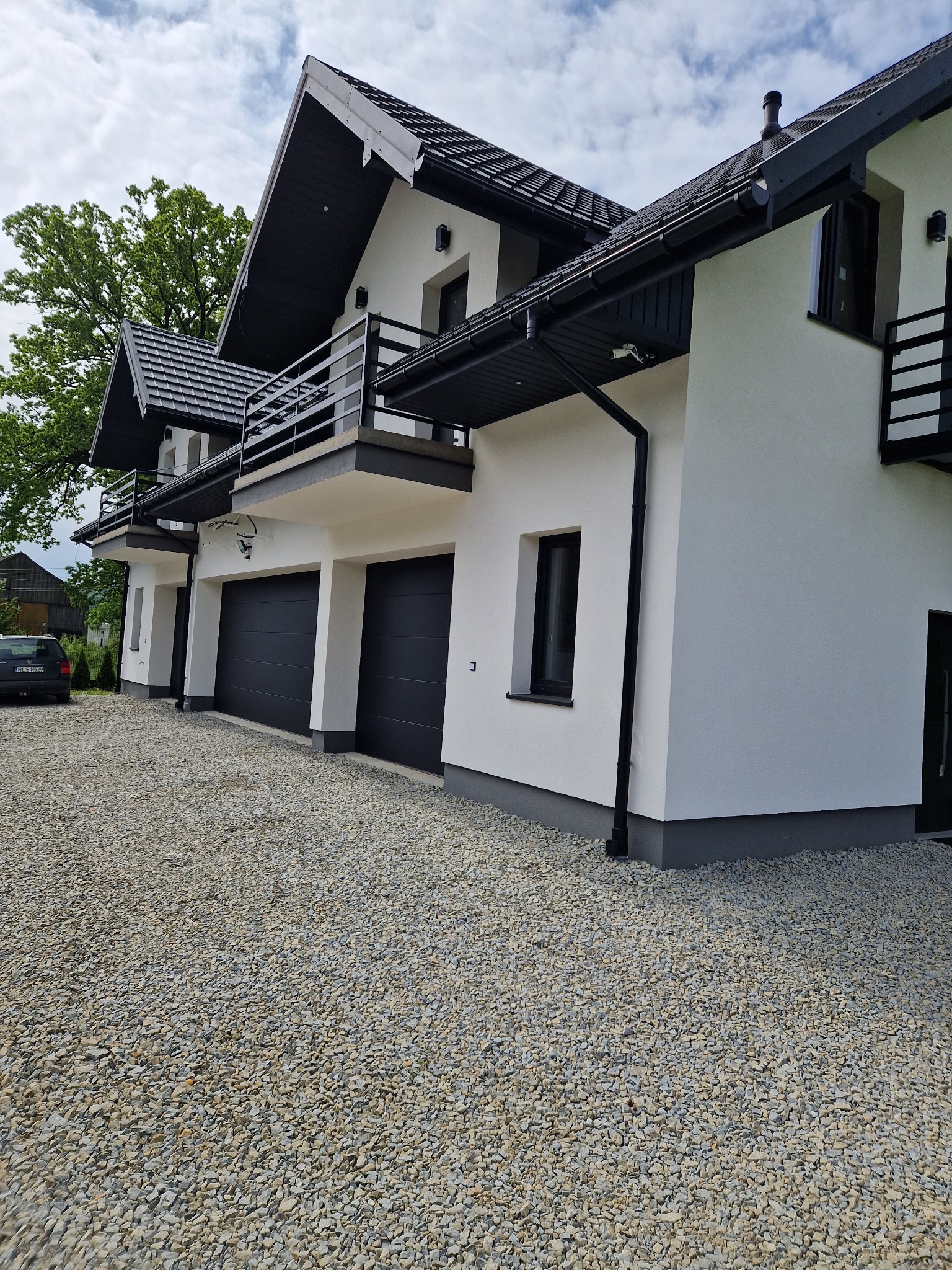 Nowoczesny, biały budynek z trzema garażami i balkonami z czarnymi elementami wykończeniowymi, otoczony żwirowym podjazdem i zielenią.