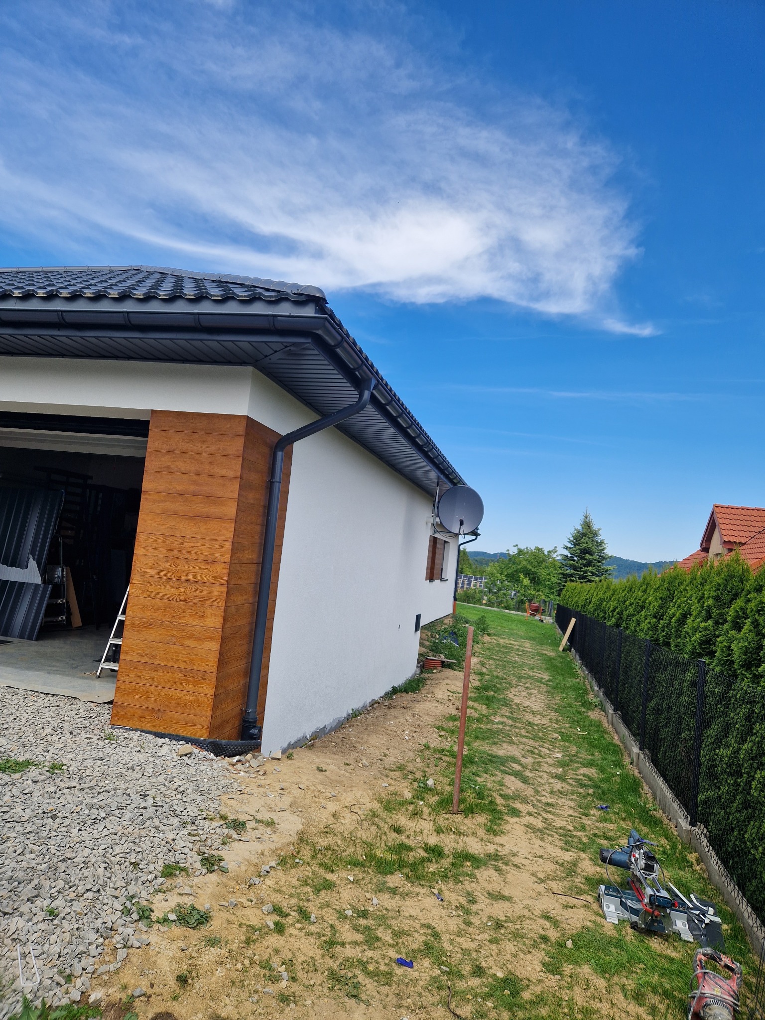 Boczny widok domu z częściowo wykończoną elewacją z paneli drewnopodobnych, narzędzia budowlane leżą na trawie obok ścieżki.