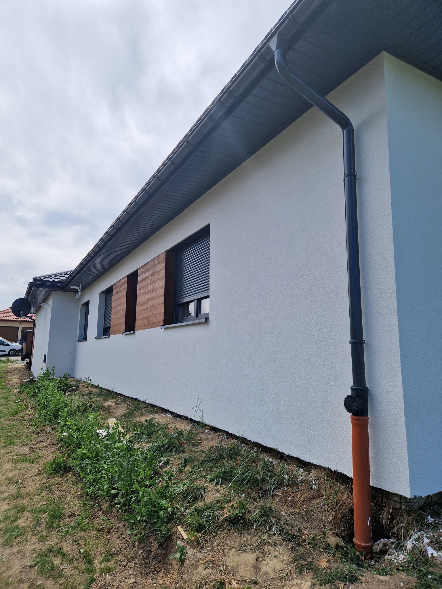 Świeżo otynkowana elewacja domu jednorodzinnego z szarymi rynnami i drewnopodobnymi panelami okiennymi, widok z boku na tle pochmurnego nieba.