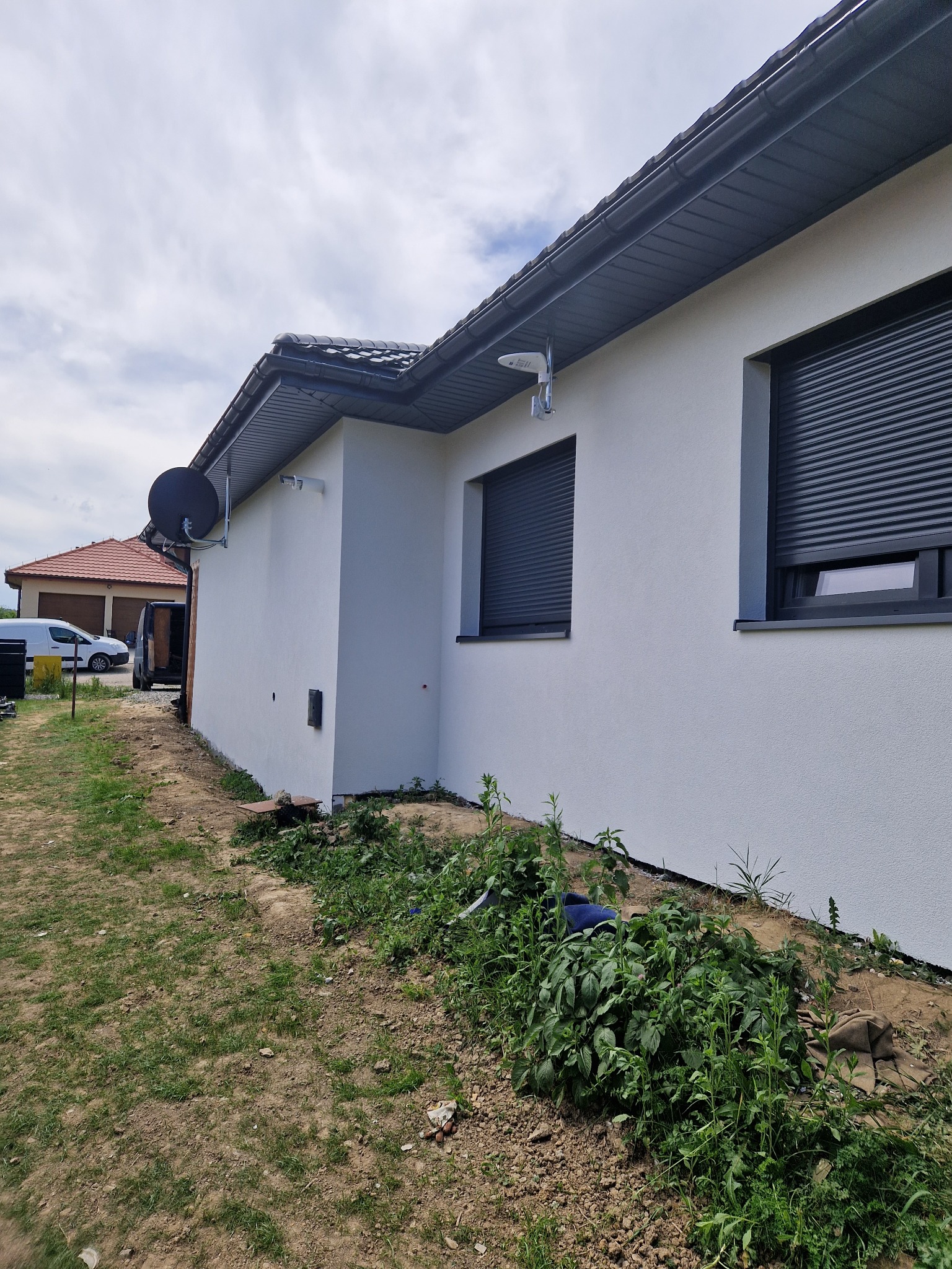 Boczny widok nowo wykończonej, białej elewacji domu z szarymi roletami okiennymi, anteną satelitarną i oświetleniem zewnętrznym, na tle zachmurzonego nieba i częściowo widocznego sąsiedniego budynku.