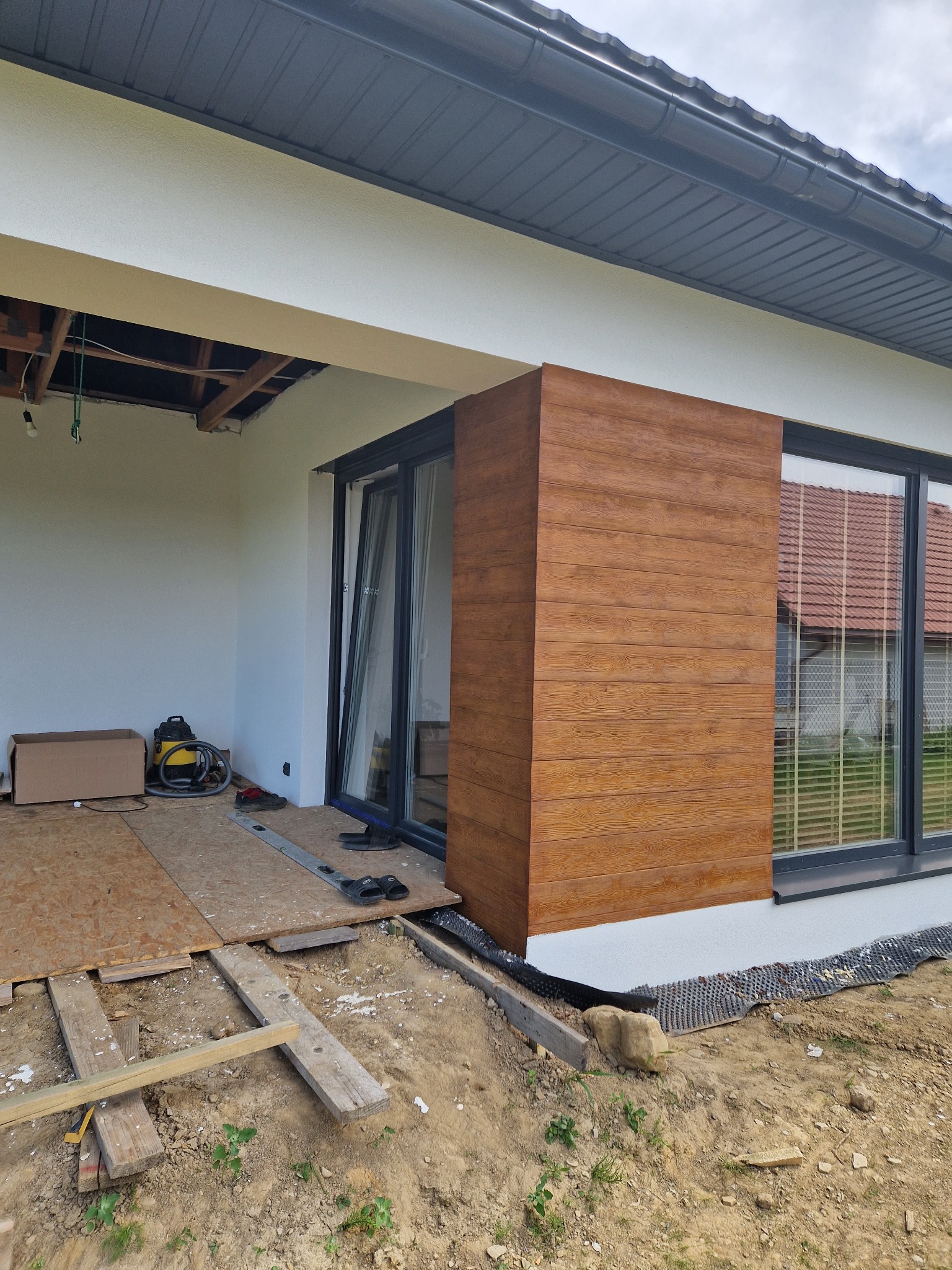 Montaż drewnopodobnej elewacji na fragmencie budynku z widocznym oknem i niezabezpieczonym stropem tarasu, na pierwszym planie ziemia i resztki materiałów budowlanych.