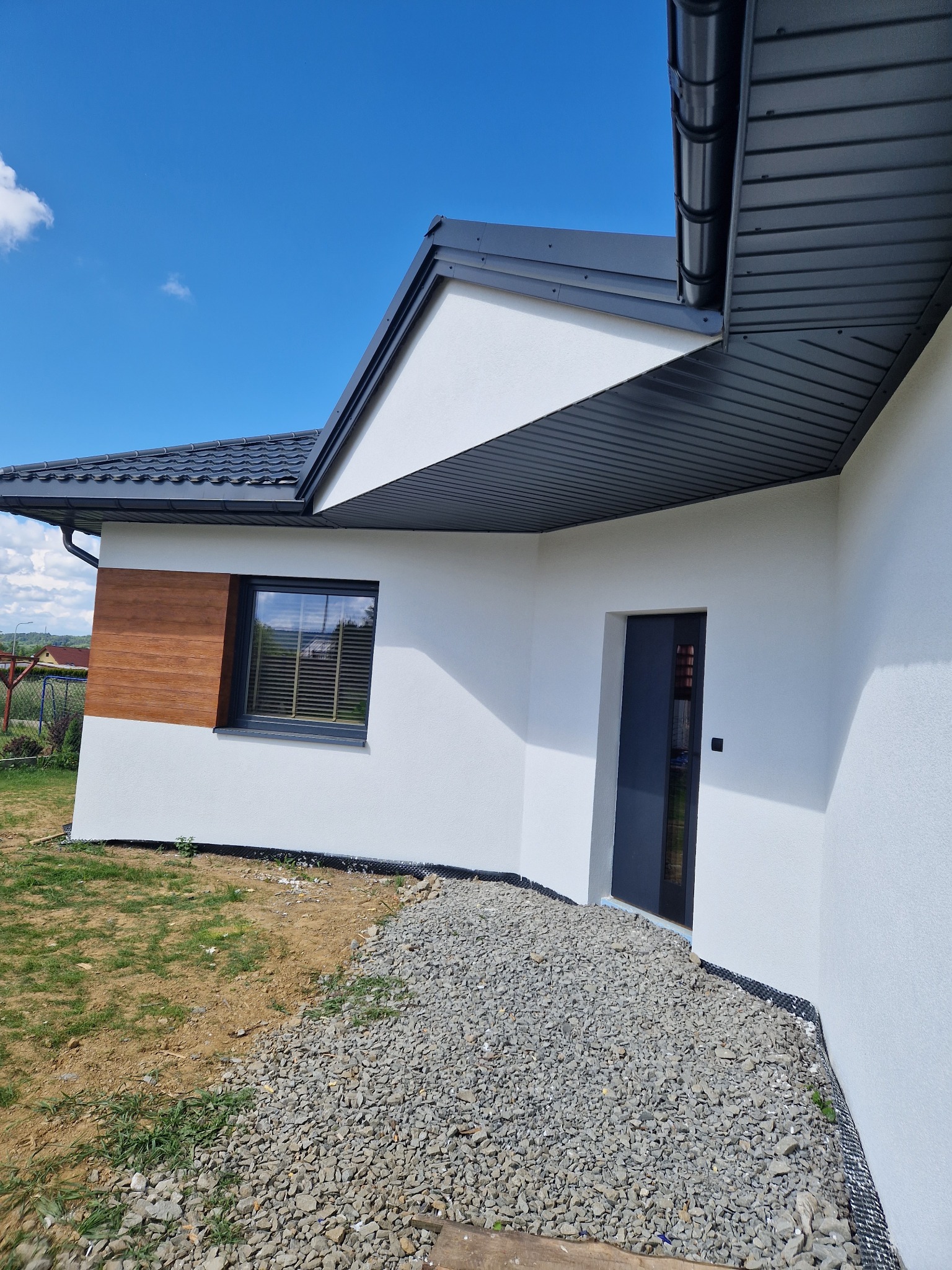 Nowoczesny dom z białą elewacją, drewnianym akcentem obok okna i szarą stolarką okienną i drzwiową, otoczony kamiennym podłożem i trawnikiem w słoneczny dzień.