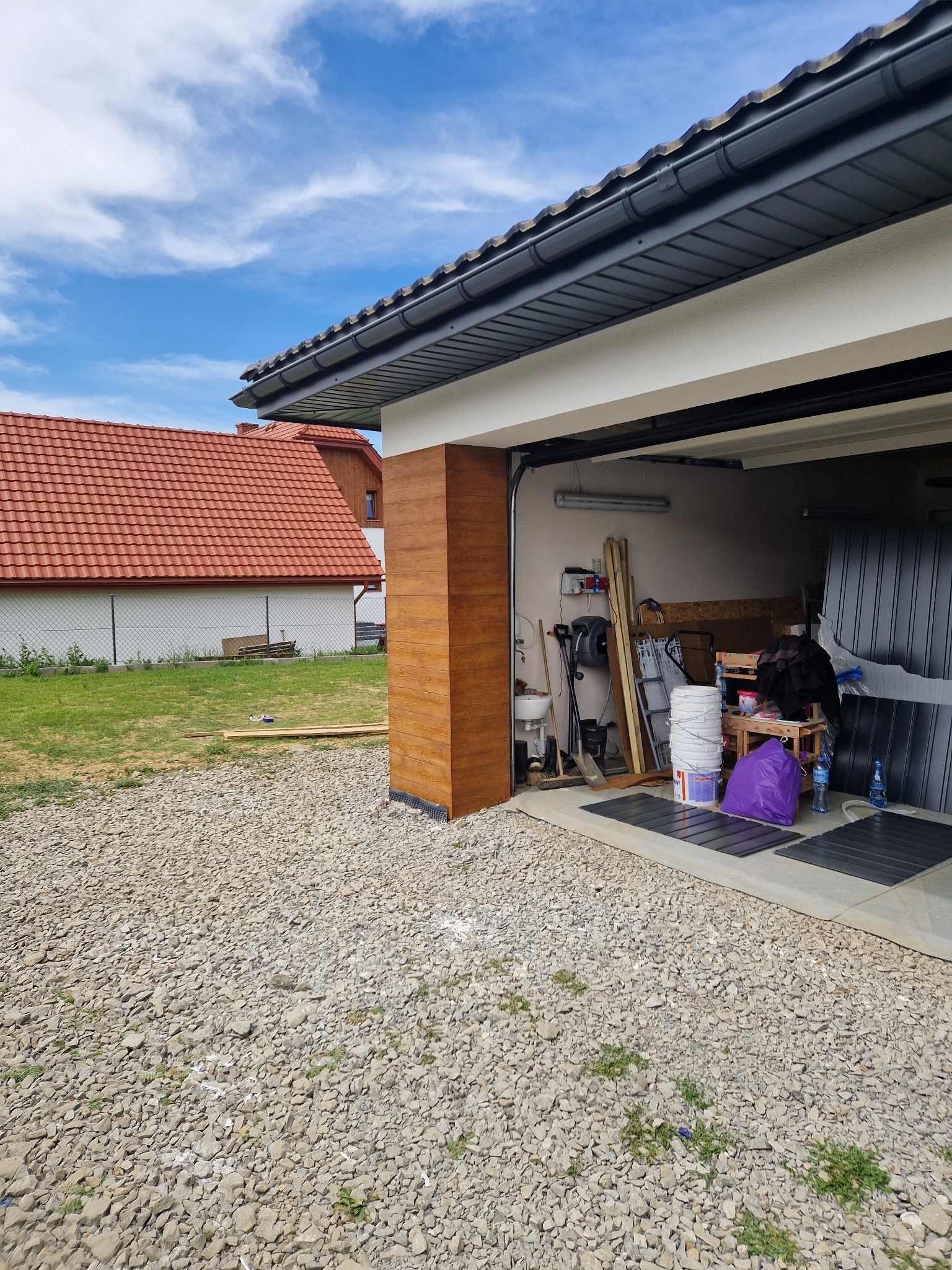 Wykończony fragment elewacji imitujący drewno przy otwartym garażu z widocznym wnętrzem zawierającym narzędzia i materiały budowlane, z domem z czerwoną dachówką w tle.