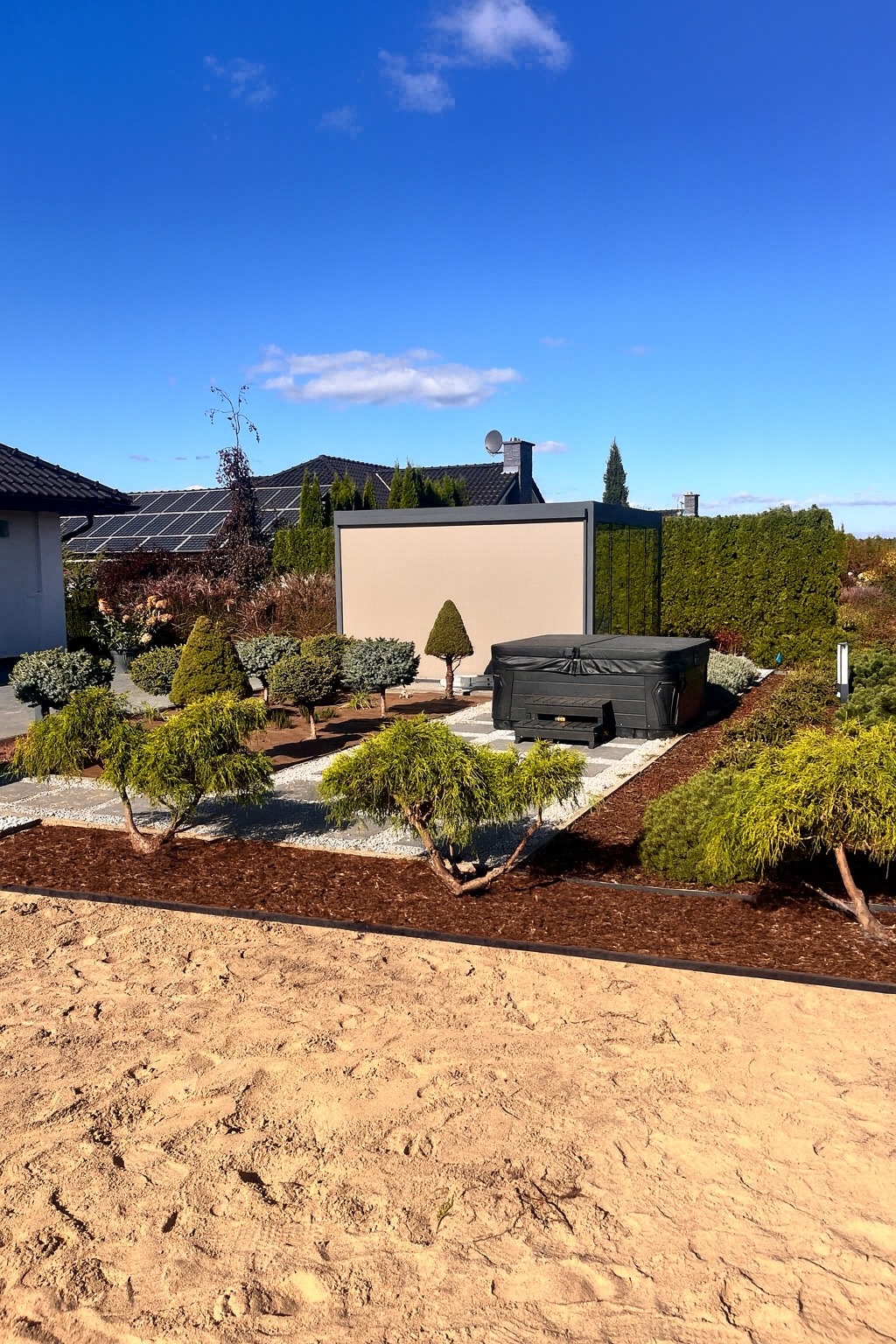 Nowoczesny ogród z jacuzzi, ozdobnymi krzewami i korą. W tle panele słoneczne na dachu domu. Błękitne niebo.