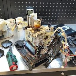 ElexFix Dawid Hłubek - Rozłożony ekspres do kawy na metalowym stole warsztatowym, widoczne elementy wewnętrzne i narzędzie. Czarny panel z otworami w tle.