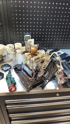 Rozłożony ekspres do kawy na metalowym stole warsztatowym, widoczne elementy wewnętrzne i narzędzie. Czarny panel z otworami w tle.