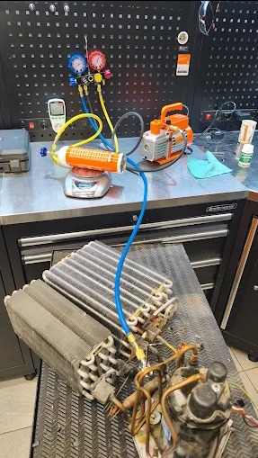 Serwis klimatyzacji: Manometry, pompa próżniowa i filtr na wadze podczas napełniania układu chłodniczego. Widoczna sprężarka i skraplacz.