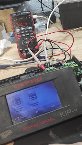 Multimetr wskazuje napięcie na panelu sterowania DEPRO KIP v4. Diagnostyka elektroniki przemysłowej w toku, widoczne przewody i złącza. Pomiar napięcia 232.88V.