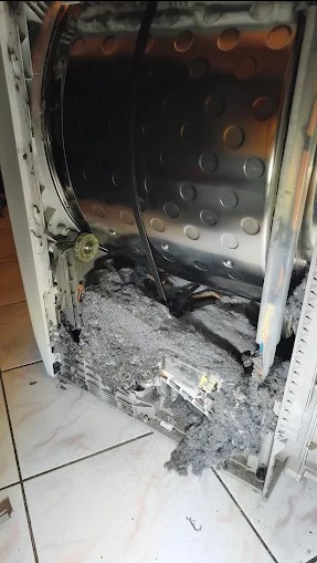 Otwarta pralka z widocznym bębnem i nagromadzeniem szarego brudu/kurzu wewnątrz obudowy. Widoczne elementy konstrukcyjne i fragment podłogi z płytek.