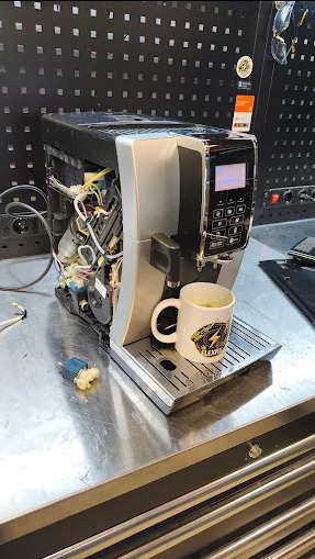 Rozłożony ekspres do kawy na metalowym stole warsztatowym, obok filiżanka z kawą. Widoczne wewnętrzne elementy urządzenia.