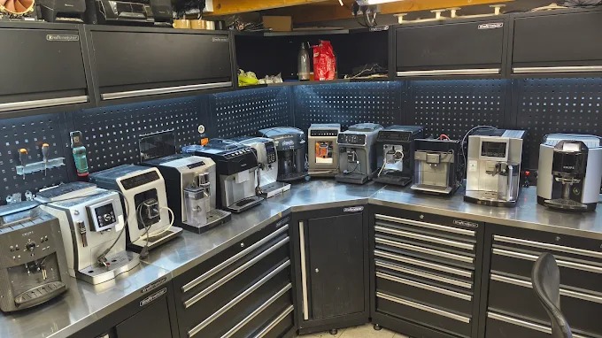 Rząd ekspresów do kawy różnych marek, ustawionych na metalowym blacie w warsztacie z narzędziami, sugerujący serwis lub naprawę. Widoczne narzędzia i akcesoria.