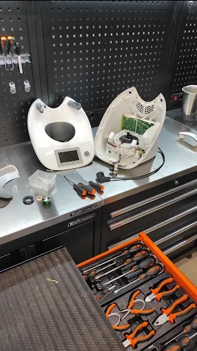 Rozłożony automat do pieczenia chleba na warsztatowym stole z narzędziami, ukazujący wnętrze urządzenia i elementy elektroniczne. Widoczny otwarty organizer z narzędziami.