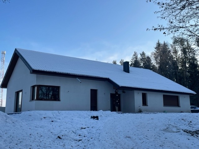 Biały dom jednorodzinny z ciemnymi oknami i dachem pokrytym śniegiem, na tle lasu i błękitnego nieba z widoczną wieżą telekomunikacyjną.