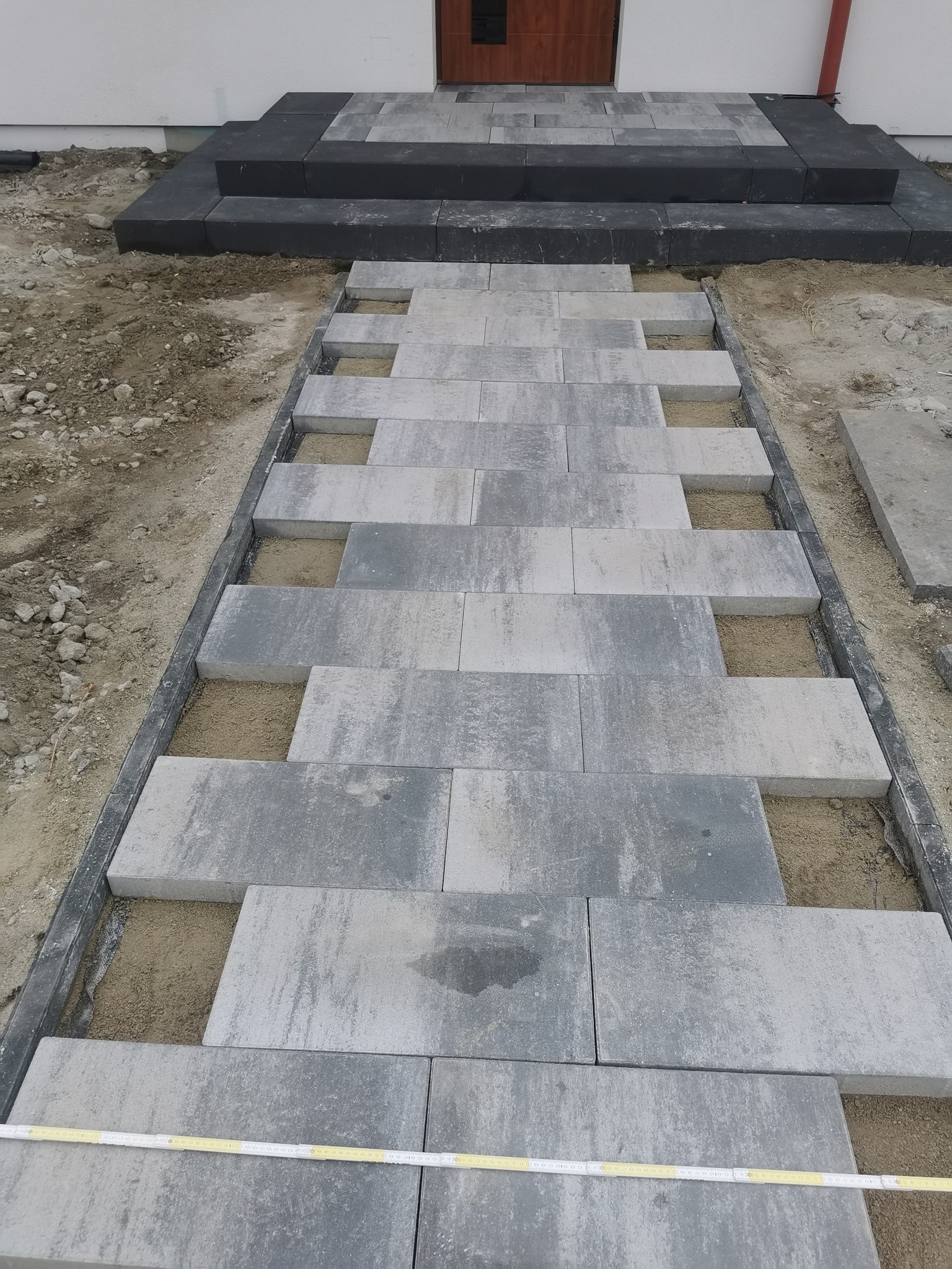 Fragment układania schodów wejściowych z szarej kostki brukowej na podsypce piaskowej. Widoczne obrzeża i częściowo ułożone stopnie.
