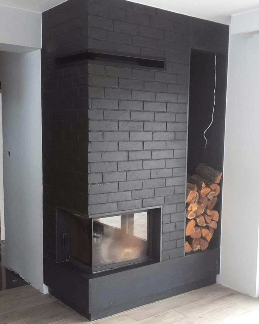 Czarny kominek z cegły z przeszklonym paleniskiem i półką na drewno, widok z boku na jasnej podłodze.