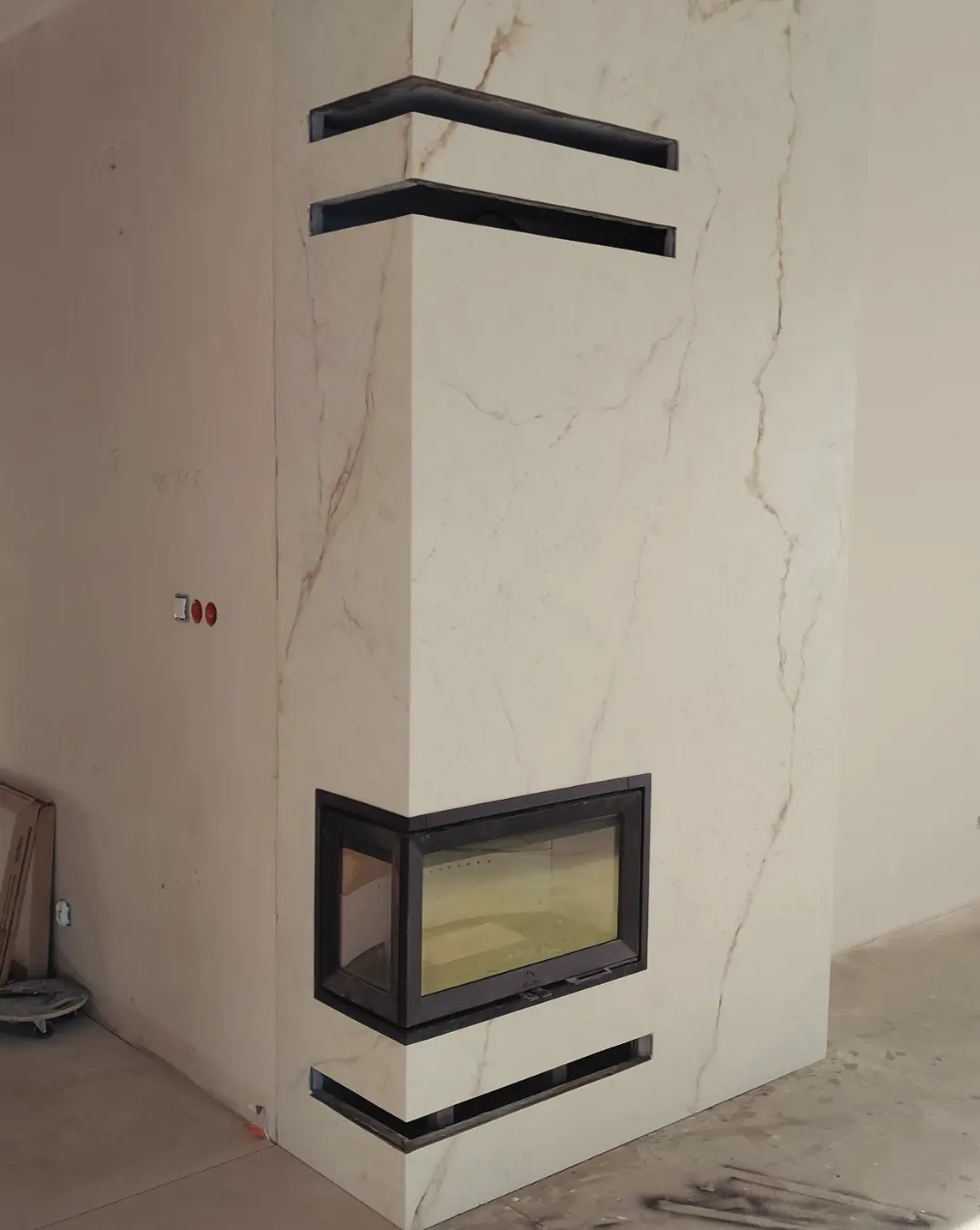 Wnętrze z narożnym kominkiem obudowanym płytami imitującymi marmur, z czarnymi, poziomymi szczelinami wentylacyjnymi, w surowym stanie wykończenia.