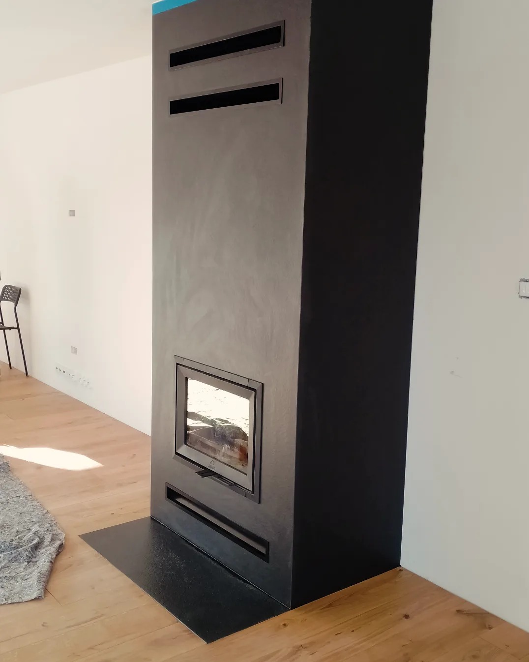 Nowoczesny, wysoki kominek w kolorze grafitowym z prostokątną szybą i czarną podstawą, umieszczony w minimalistycznym wnętrzu z drewnianą podłogą.