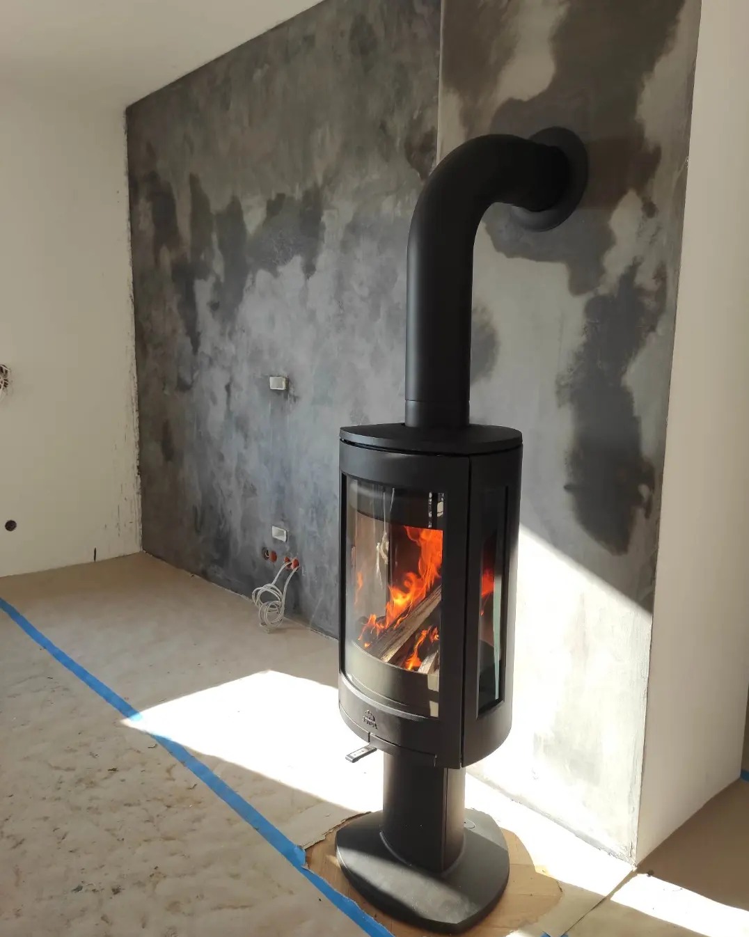 Czarny, wolnostojący piec kominkowy z płomieniami w trakcie montażu, widoczne zabezpieczenia podłogi taśmą malarską, surowa ściana z efektem betonu.