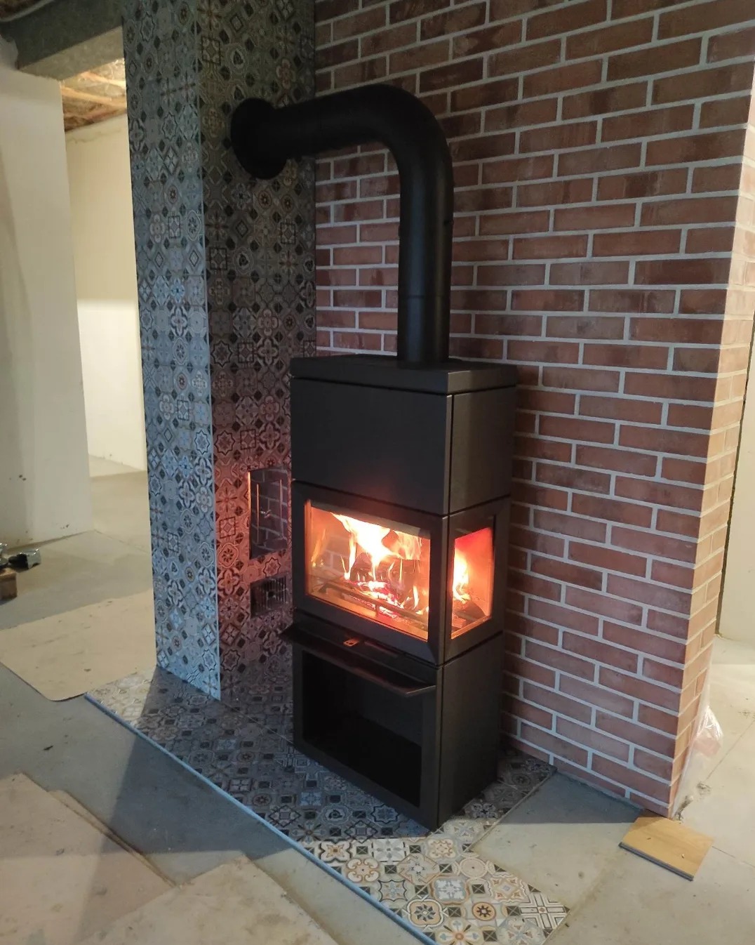 Nowoczesny, czarny piec wolnostojący z płomieniami widocznymi przez szybę, ustawiony na dekoracyjnych płytkach podłogowych, z ceglanym wykończeniem ściany w tle.