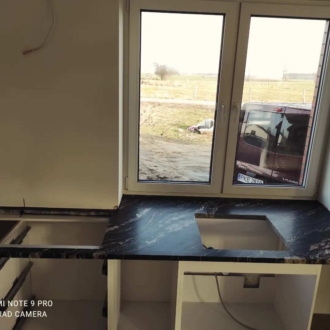 Montaż czarnego blatu kamiennego z wycięciem na zlew przy oknie w surowym wnętrzu kuchni, widoczny krajobraz za oknem i odbicie samochodu.
