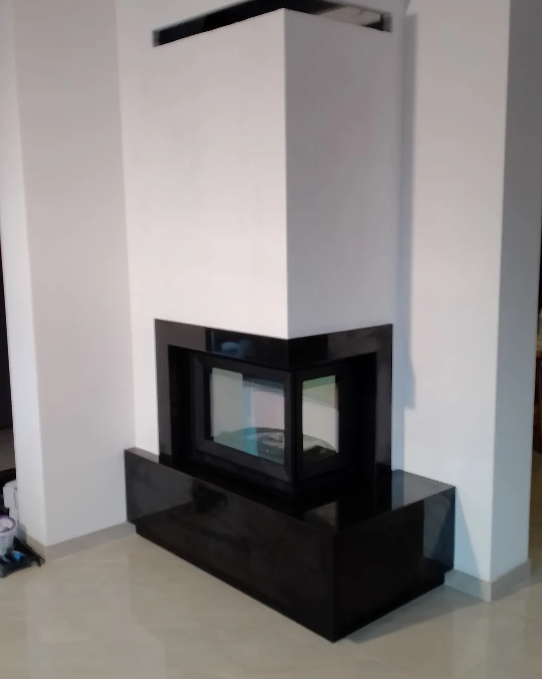 Nowoczesny kominek narożny z czarnym, błyszczącym obramowaniem i białym wykończeniem, w minimalistycznym wnętrzu.