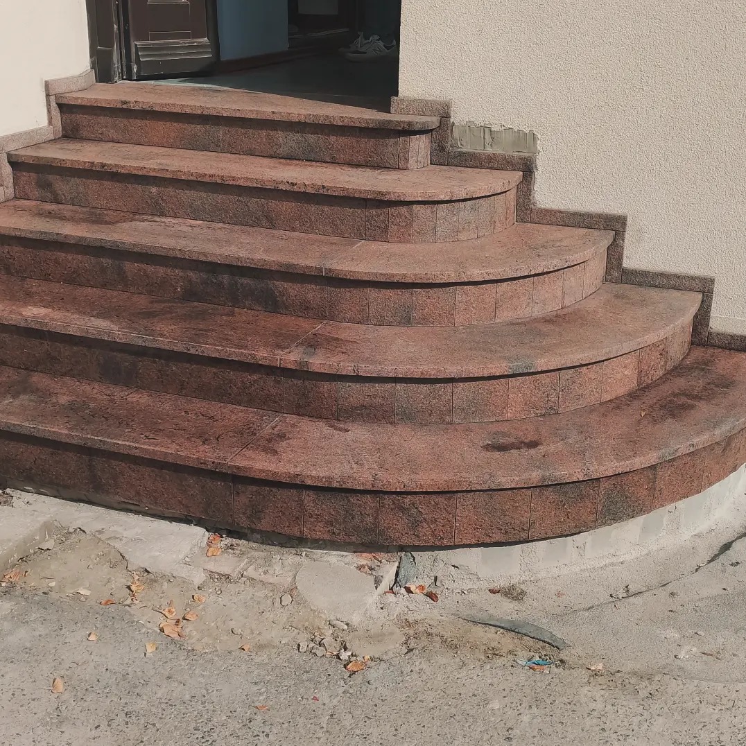 Zewnętrzne schody wejściowe wyłożone płytkami imitującymi czerwony granit, z zaokrąglonymi krawędziami stopni, widoczne ślady po pracach budowlanych.