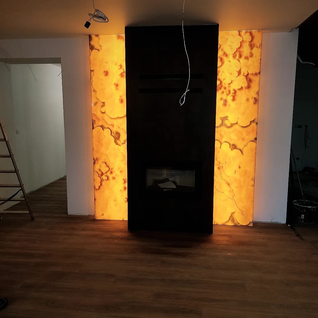 Nowoczesny kominek z czarną obudową i podświetlanymi panelami z onyksu, widoczna drabina i niedokończone ściany sugerują prace wykończeniowe.