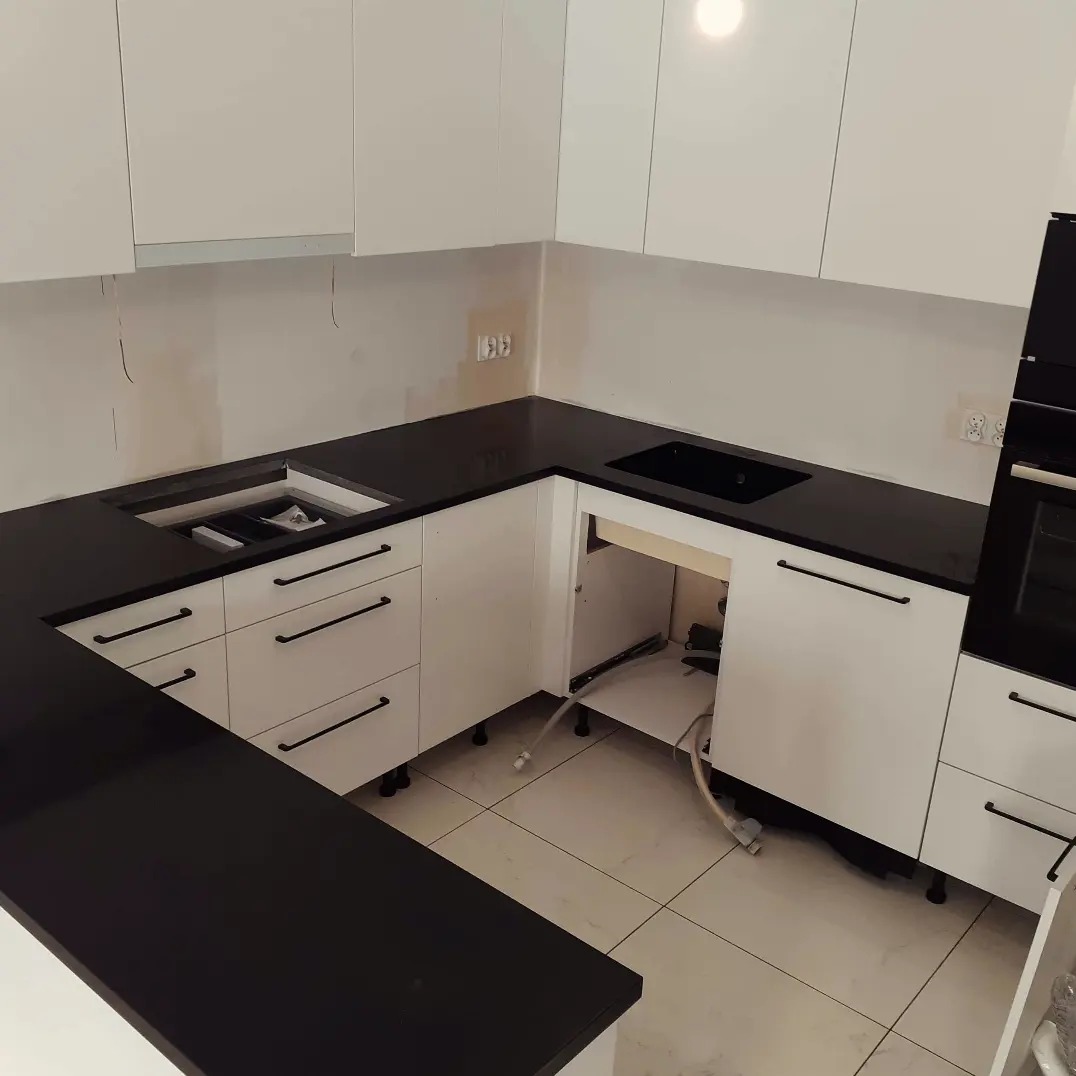 Montaż czarnego blatu kuchennego na białych szafkach z długimi, czarnymi uchwytami; widoczne niedokończone prace instalacyjne pod zlewem.