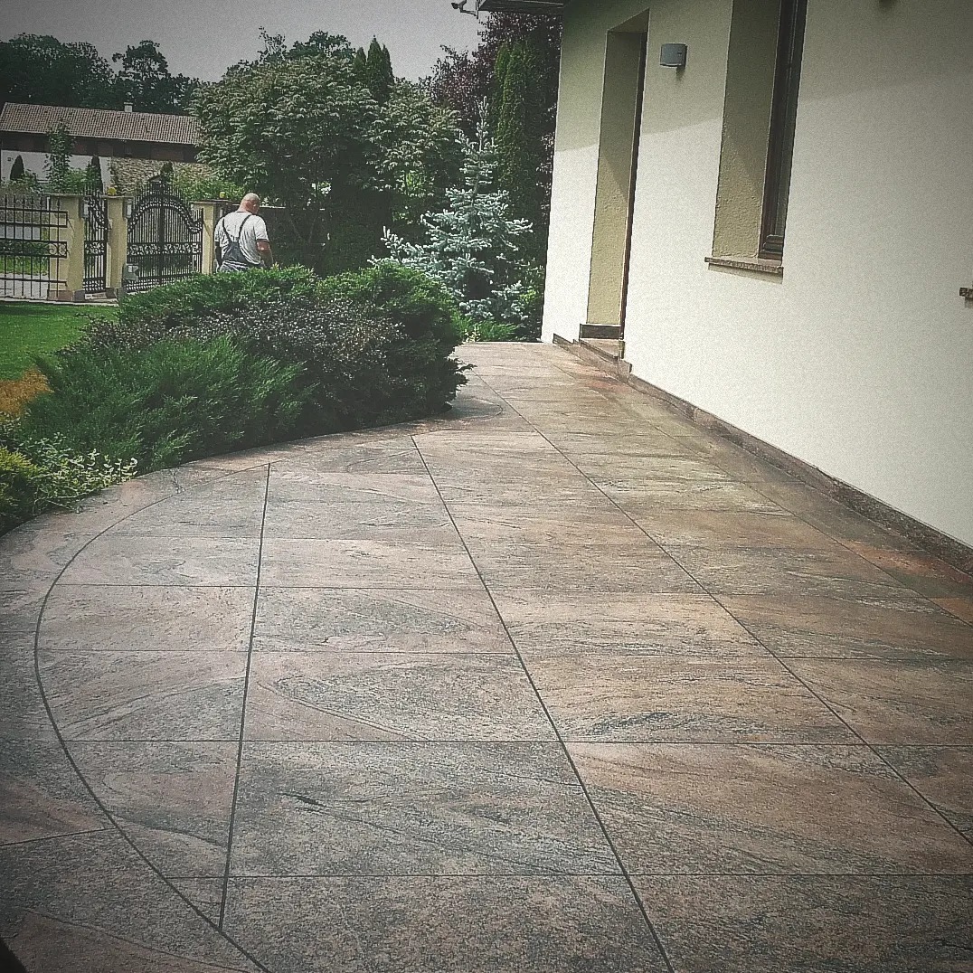 Ułożony taras z płyt kamiennych o nieregularnym wzorze, z zaokrąglonym brzegiem obok krzewu, w tle widoczny mężczyzna w ogrodzie.